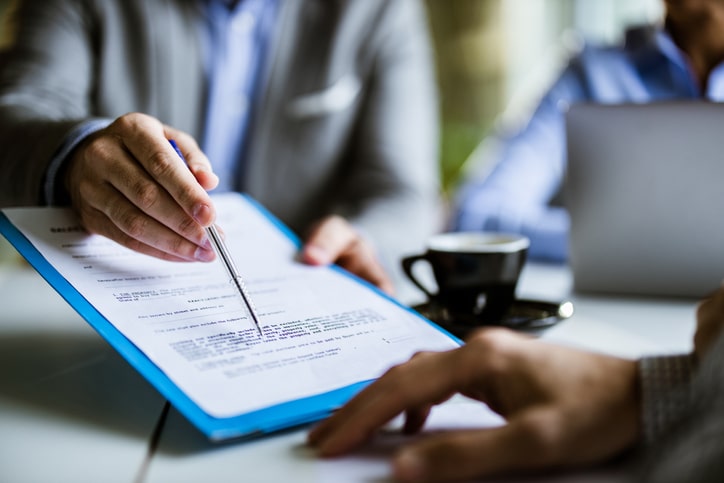 Two individuals in a business setting review and discuss a document on a clipboard. One person is pointing to a section with a pen, while the other is holding the clipboard. A cup of coffee is visible. They are franchise consultants discussing potential strategies for growth with great focus and collaboration.