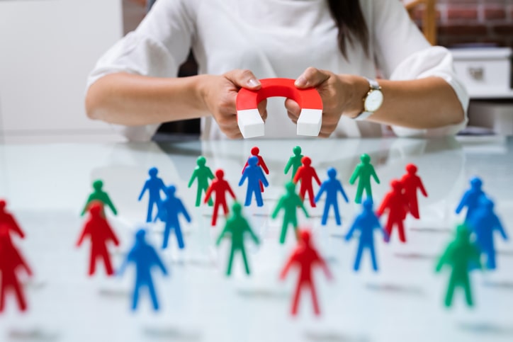 A person holds a large magnet attracting red, blue, and green human-shaped figures on a table, demonstrating the magnetic pull similar to how franchise consultants in Australia attract potential business partners.