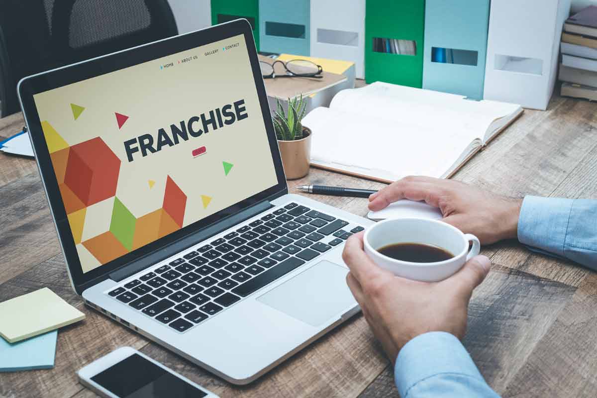 A person holding a coffee cup sits at a desk with an open laptop displaying the word "FRANCHISE". Office supplies and a plant are on the desk, suggesting they might be receiving advice from franchise consultants.