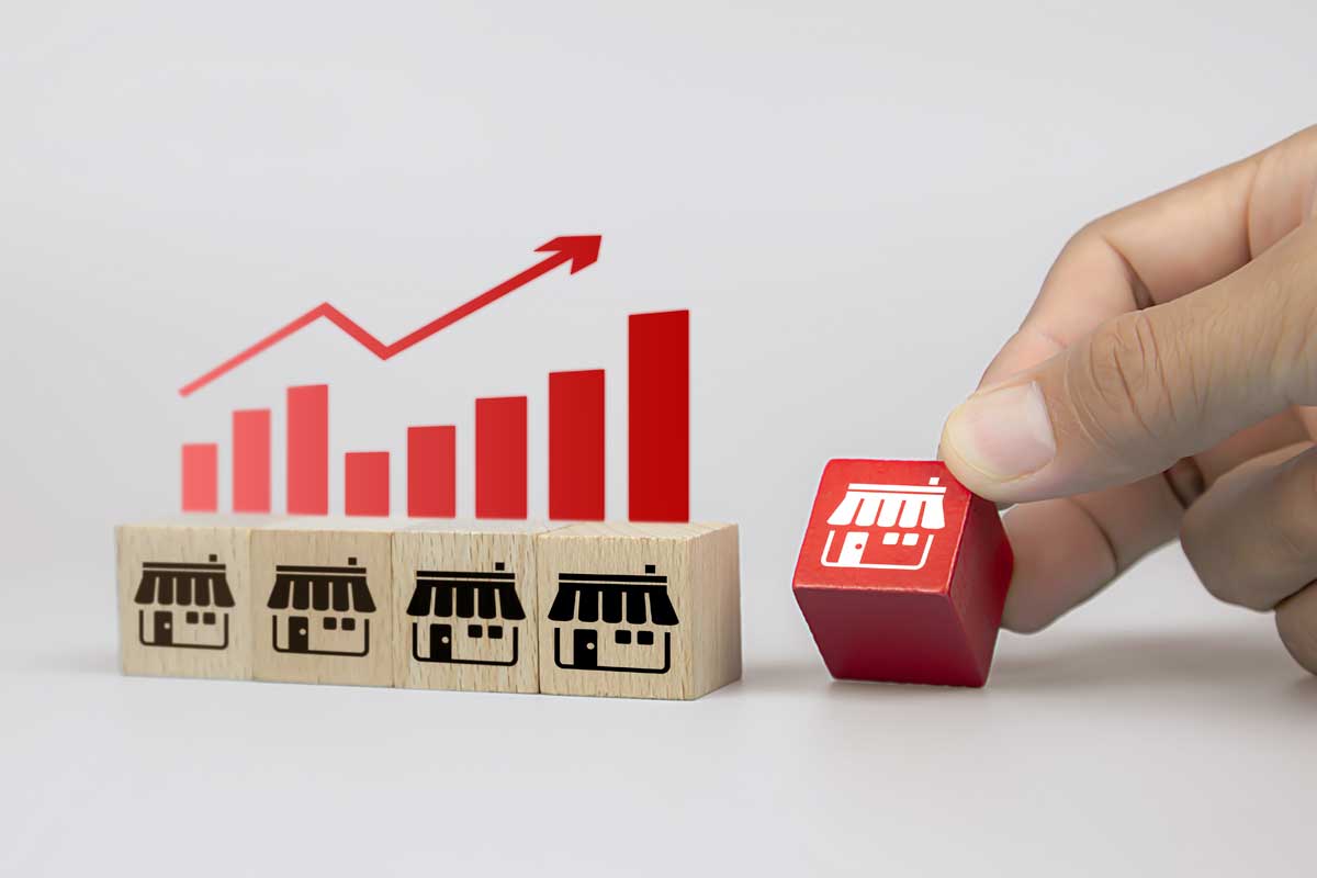 A hand arranges a red building block with a storefront symbol next to three wooden blocks bearing the same mark, while an upward trend graph is visible. This scene represents the expert strategy of franchise consultants in Australia.