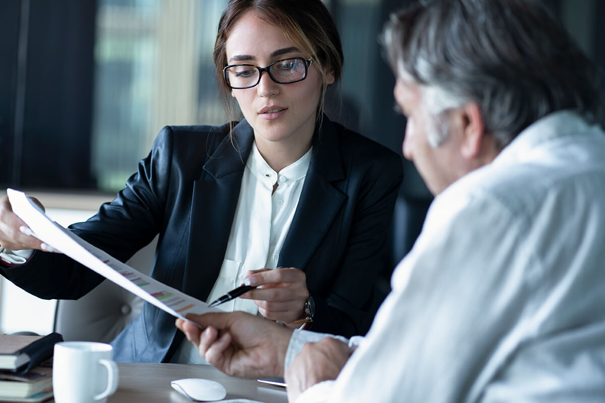 A woman in a business suit, holding a pen, discusses a document with a man in a white shirt at an office table with a coffee cup and computer mouse. The meeting appears to involve the expertise of franchise consultants from Australia.