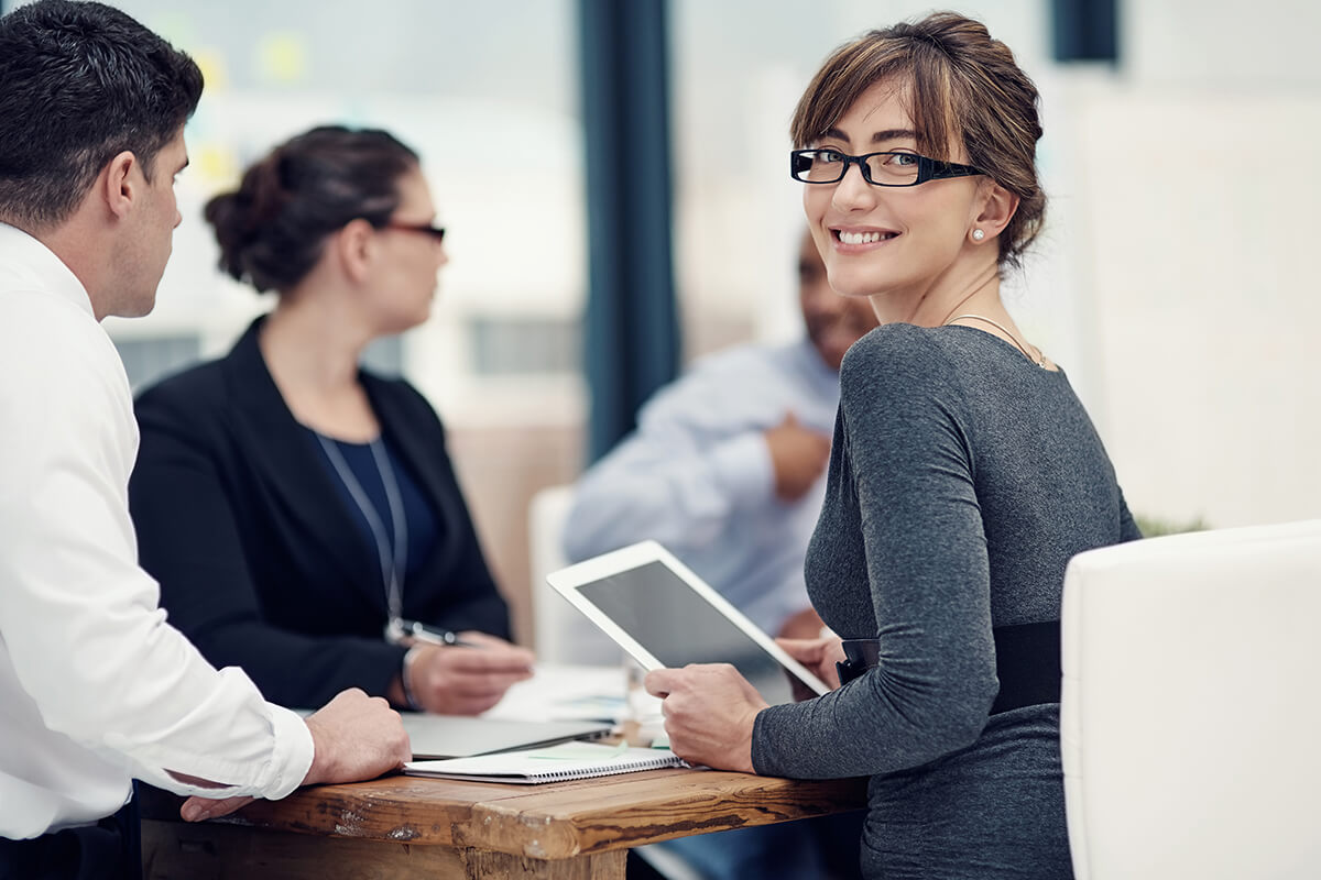 A woman holding a tablet smiles at the camera while sitting at a table with three colleagues, who are franchise consultants engaged in discussion.