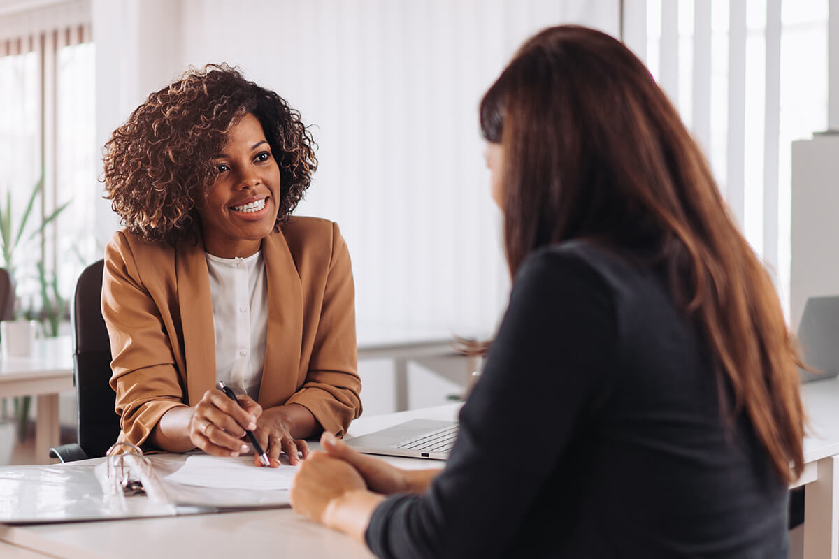 Two women in a meeting, seated at a desk. One woman is holding a pen and speaking, discussing strategies with the other woman, who listens attentively. Documents and a laptop are on the desk as they consult on franchise business opportunities in Australia.