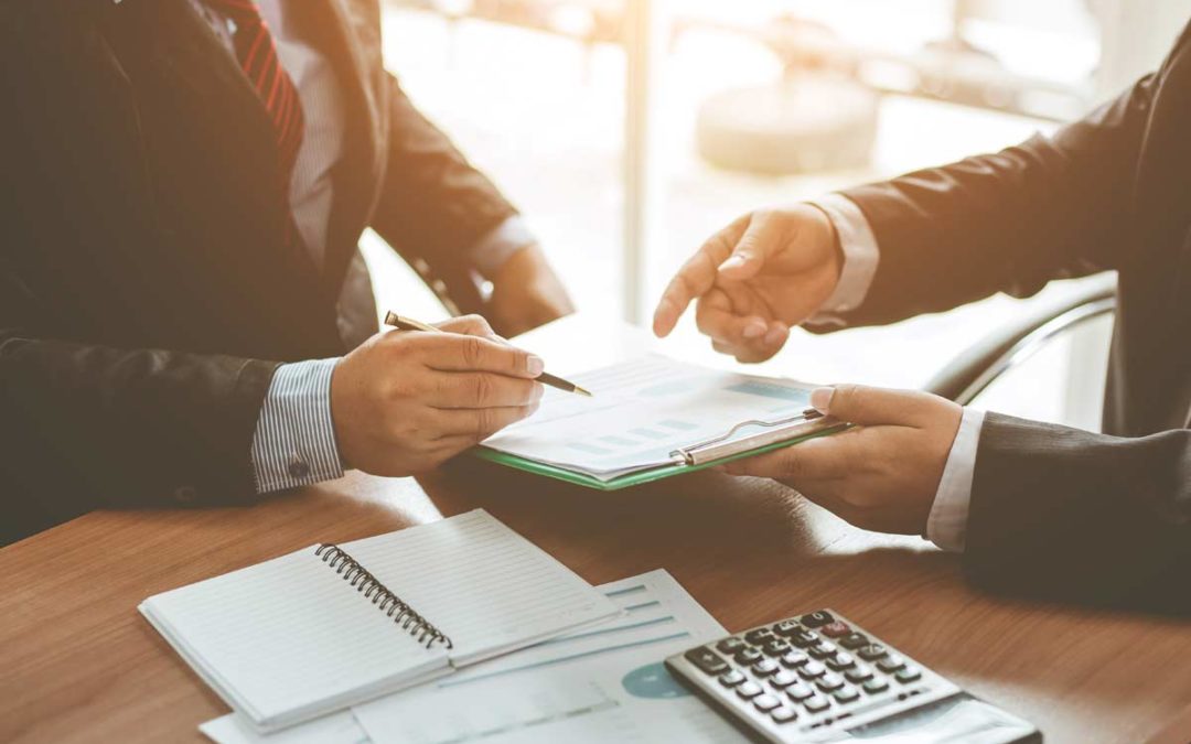 Two people in business attire are discussing a document with charts at a wooden table, which also has a notebook, pen, and calculator. One person, likely a franchise consultant, is holding a pen and pointing at the document.