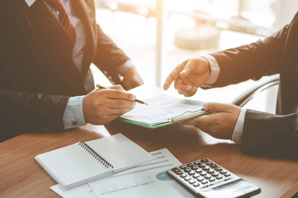 Two people in business attire are discussing a document with charts at a wooden table, which also has a notebook, pen, and calculator. One person, likely a franchise consultant, is holding a pen and pointing at the document.