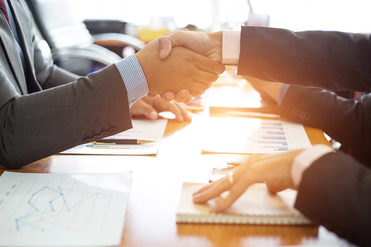 Two individuals in suits shake hands across a desk cluttered with documents, charts, and stationery, indicating a business agreement or partnership. One could be a franchise business consultant assisting the other in navigating the complexities of expanding their operations.