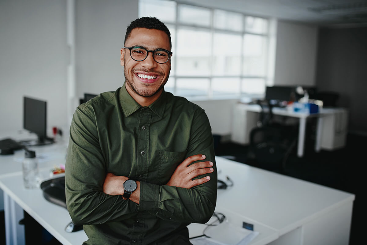 A person wearing glasses and a green shirt stands smiling in an office setting with arms crossed. Office desks and computers are visible. They work as franchise consultants, assisting businesses with expert advice.